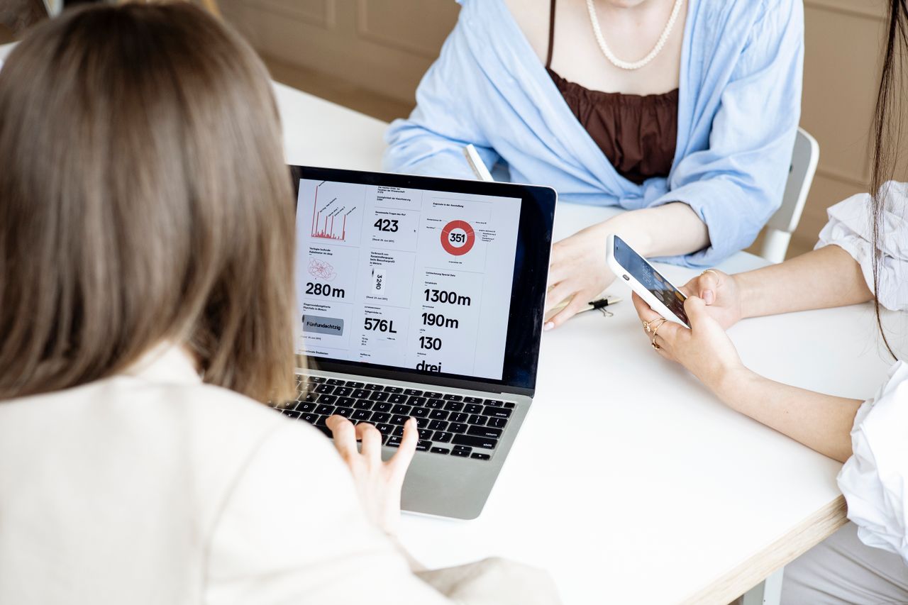 A group of women collaborating at a desk using a laptop and smartphone for business tasks indoors.